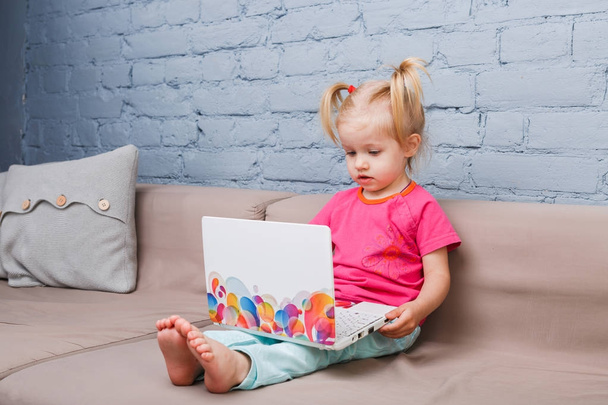 dziecko dziewczynka trzech lat używa szkolenia laptopa siedząc na kanapie w pomieszczeniu z niebieski ceglany mur. Dziecko o niebieskich oczach i blond z śmieszne ogony na głowie jest ubrana w jasne jerse - Zdjęcie, obraz