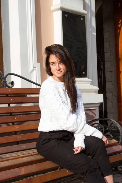 Young girl in a white sweater sits on a bench on a building background - Foto, Imagem