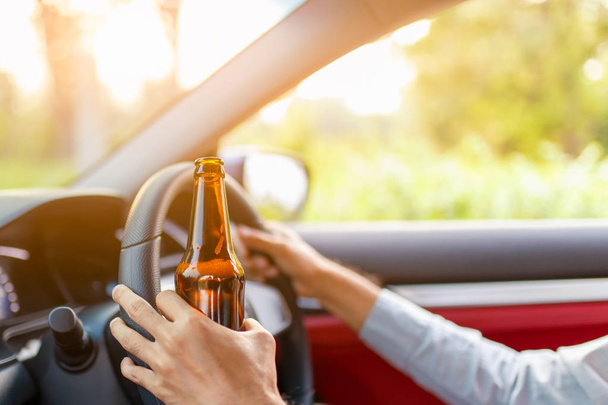 Drunk young man drives a car with a bottle of beer with sunset background, Dangerous driving concept - Photo, Image