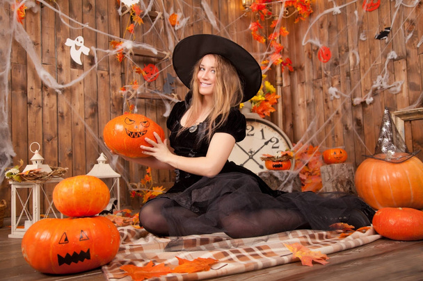 beautiful classic witch woman wearing black sorceress velvet dress and pointed hat, with orange evil halloween pumpkins and spiderweb  - Photo, Image