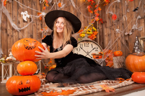 beautiful classic witch woman wearing black sorceress velvet dress and pointed hat, with orange evil halloween pumpkins and spiderweb  - Photo, Image