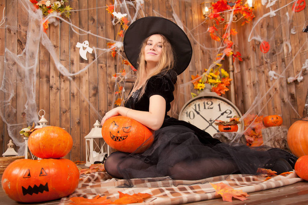 gyönyörű klasszikus boszorkány nő boszorkány fekete bársony ruhát visel, és rámutatott a kalap, narancssárga gonosz halloween sütőtök és spiderweb  - Fotó, kép