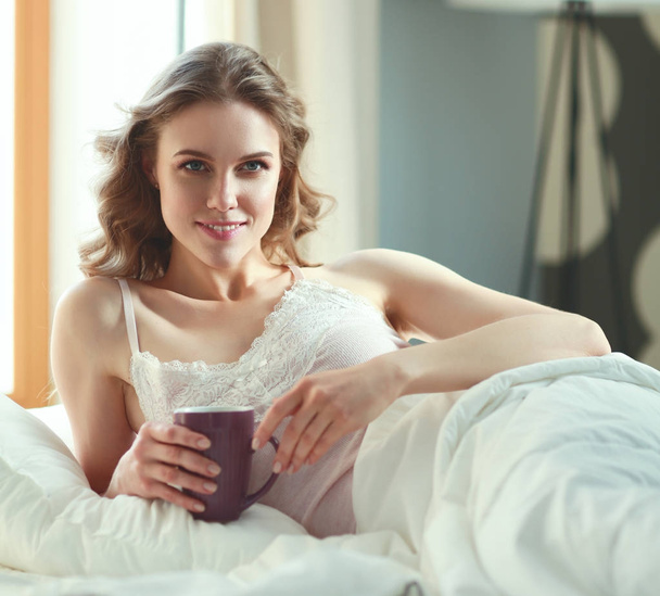 Young woman drinking cup of coffee or tea while lying in bed - Foto, Imagen