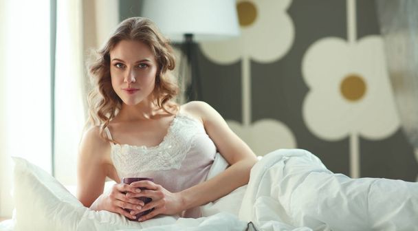 Young woman drinking cup of coffee or tea while lying in bed - Фото, зображення