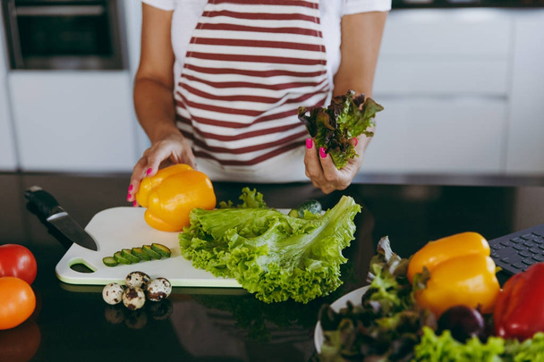 Young happy woman holding vegetables in hands in kitchen with laptop on the table. Vegetable salad. Dieting concept. Healthy lifestyle. Cooking at home. Prepare food Close up. - Foto, Imagen