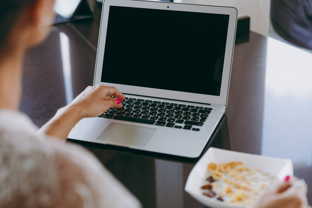 Jovem mulher trabalhando com laptop enquanto café da manhã com cereais e leite. Comer em casa de perto. Vista traseira. Com lugar para texto
. - Foto, Imagem