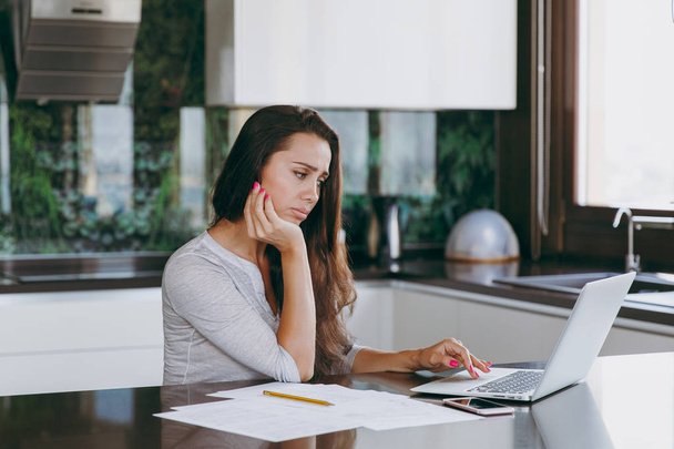 Atraente mulher de negócios pensativa e triste trabalhando com documentos e laptop na cozinha em casa
 - Foto, Imagem
