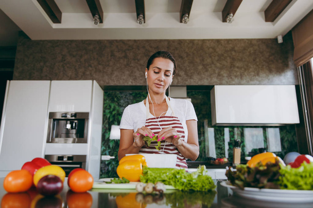 Jeune femme avec des écouteurs dans les oreilles tenant des légumes dans les mains dans la cuisine avec un ordinateur portable sur la table. Salade de légumes. Concept de régime. Mode de vie sain. Cuisiner à la maison. Préparer les aliments
 - Photo, image