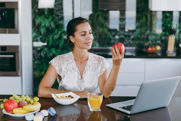 Retrato de la hermosa mujer feliz que trabaja con el ordenador portátil, mientras que el desayuno con cereales y leche. Comer en casa
. - Foto, Imagen
