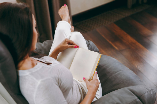 Молодая женщина сидит дома на современном стуле перед окном, отдыхает в гостиной и читает книгу. Вид сверху
. - Фото, изображение