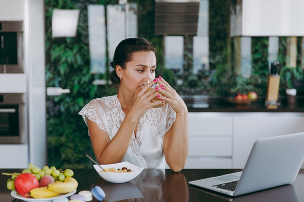 Retrato de una hermosa mujer joven que trabaja con el ordenador portátil, mientras que el desayuno con cereales y leche y beber jugo de naranja. Comer en casa
. - Foto, imagen