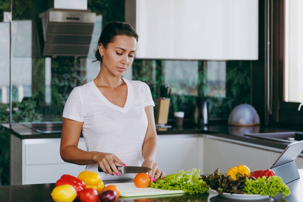 Una joven corta verduras en la cocina con un cuchillo y un portátil sobre la mesa. Ensalada de verduras. Concepto de dieta. Estilo de vida saludable. Cocinar en casa. Prepara comida. Con lugar para el texto
 - Foto, Imagen