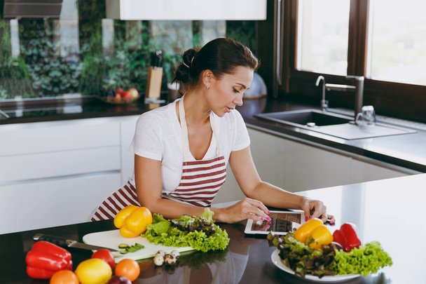 Young happy woman in apron looking at recipe in tablet in the kitchen. Vegetable salad. Dieting concept. Healthy lifestyle. Cooking at home. Prepare food. - 写真・画像