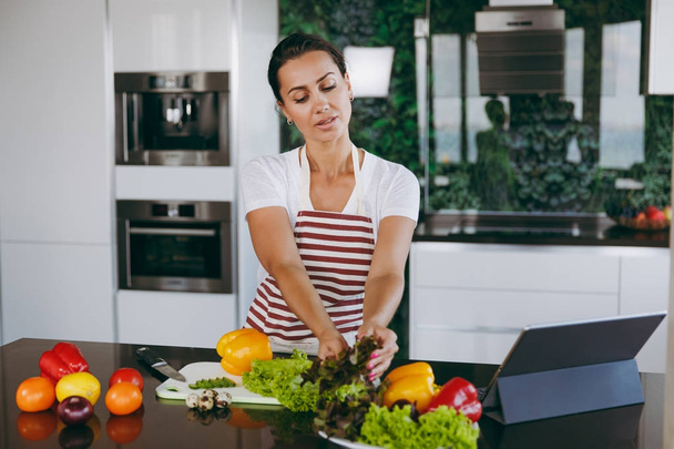 Young happy woman holding vegetables in hands in kitchen with laptop on the table. Vegetable salad. Dieting concept. Healthy lifestyle. Cooking at home. Prepare food. - Fotoğraf, Görsel