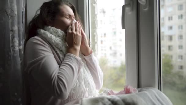 Una donna fredda e fredda starnutisce. Ragazza soffiando il naso mentre seduto alla finestra
. - Filmati, video