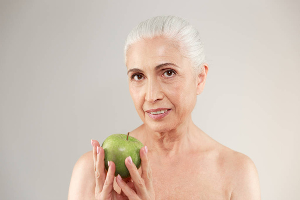Concentrated naked elderly woman holding apple. - Foto, afbeelding