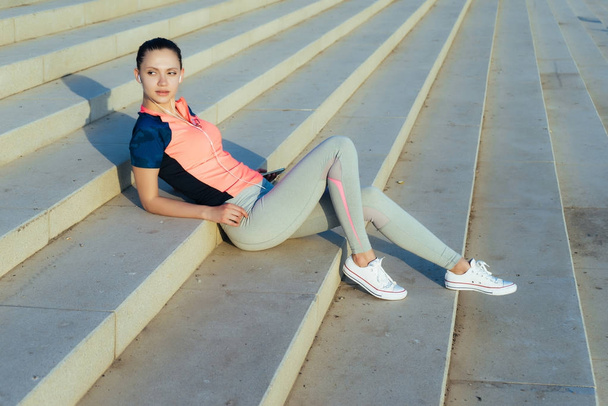 young sporty beautiful girl resting on the steps in the rays of the sun - Фото, изображение
