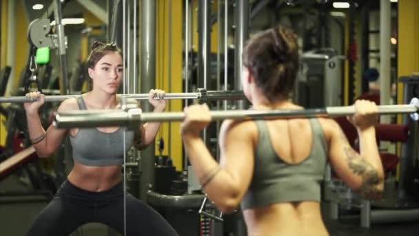 La joven deportista está haciendo sentadillas con una barra en un gimnasio frente a un espejo
 - Metraje, vídeo