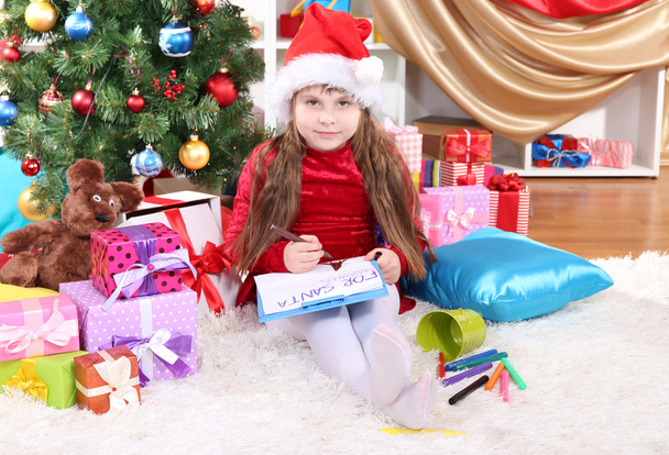 Beautiful little girl in red dress writes letter to Santa Claus in festively decorated room - Φωτογραφία, εικόνα