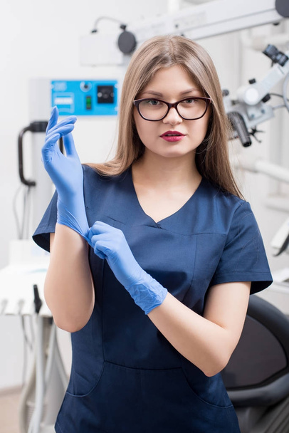 Portrét mladé ženy zubař v zubní ordinaci morden. Doktor nosíš brýle, modré uniformě, modré rukavice. Zubní lékařství - Fotografie, Obrázek