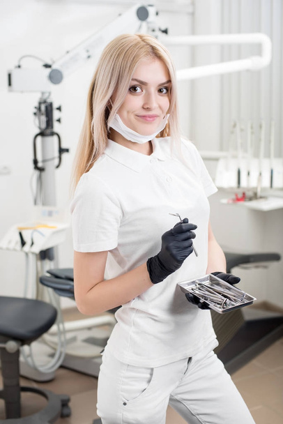 Портрет молодой красивой женщины-стоматолога, держащей стоматологический инструмент в морденском стоматологическом кабинете. Доктор в черных перчатках и смотрит в камеру. Стоматология
 - Фото, изображение