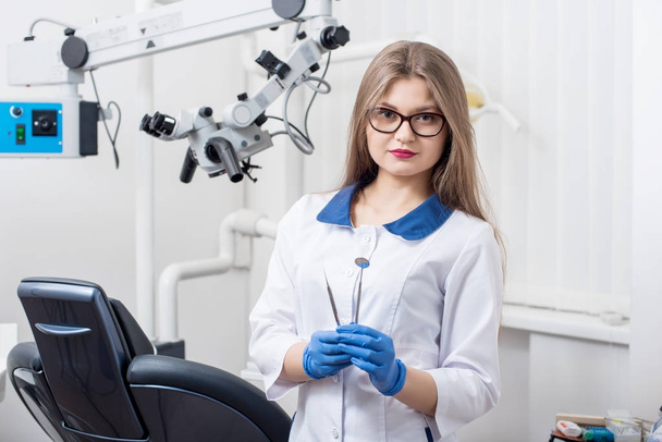 Portrét mladé krásné ženské zubař držící zubní nástroje v morden zubní ordinaci. Doktor nosí brýle, bílé dresy, modré rukavice a při pohledu do kamery. Zubní lékařství - Fotografie, Obrázek