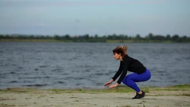Beautiful sports girl does burpees jumping on the beach near the flowing river. - Footage, Video