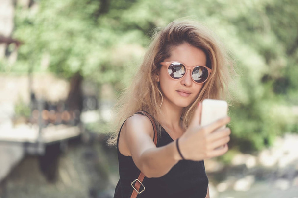 Closeup portrait of young beautiful woman in sunglasses with long blonde curly hair doing selfie photos - Foto, afbeelding