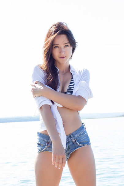 Girl on the beach in a white shirt and short denim shorts - 写真・画像