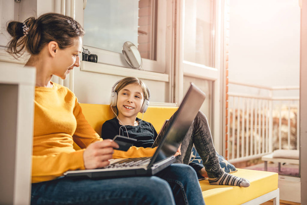 Mother and daughter sitting on yellow sofa and talking while using laptop and tablet - Фото, изображение
