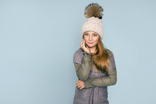 Schattig klein meisje gekleed in winter kleding, hoed en winter jas met bont, permanent op blauwe achtergrond - Foto, afbeelding