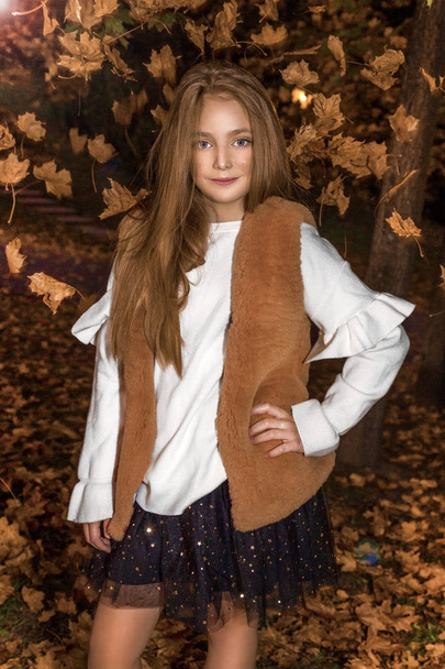 Carina bambina vestita con cappotto e stivali seduta su sfondo autunnale, foglie d'autunno dorate
. - Foto, immagini