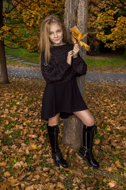 Милая маленькая девочка, одетая в пальто и сапоги сидя на осеннем фоне, золотые осенние листья
. - Фото, изображение
