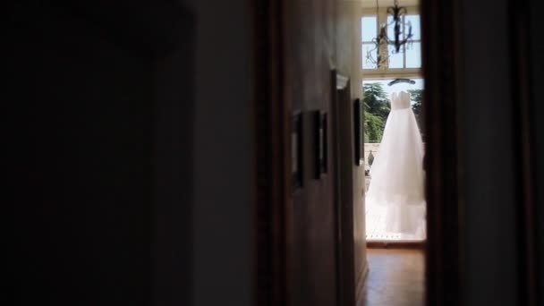 Witte jurk hangen in deuropening van balkon licht te wachten voor slijtage. Lichte witte avond of trouwjurk op hanger gezien vanaf donkere gang kamer. Bruids mode ontwerp ochtend bruid zomer garderobe luchten - Video