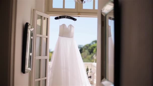 White dress hangs on special hanger in doorway to terrace close up. Light summer gown made of silk or chiffon fabrics waits for bride at sunny morning balcony. Wedding fashion tailoring design studio - Footage, Video