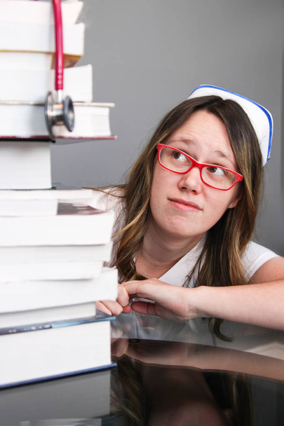 Молодая медсестра в кепке смотрит на стопку книг
 - Фото, изображение