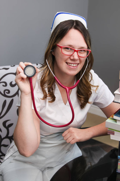Jovem enfermeira com tampa segurando estetoscópio e sorrindo
 - Foto, Imagem
