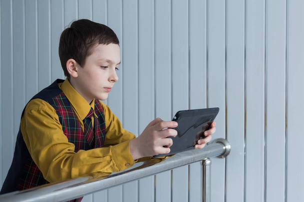 A young guy with a serious face in a mustard shirt and a checkered waistcoat looks at the black tablet on the background of the striped wall. - Photo, Image