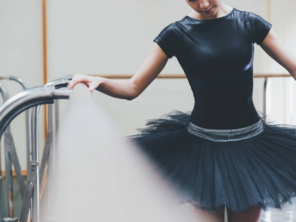 Ballerina in black tutu and pointe stretches on barre in ballet gym. Woman standing near bar and mirror, preparing for perfomance. - Photo, Image