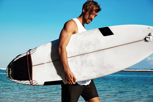 Ritratto di bel hipster abbronzato moda uomo modello surfista indossa abiti casual andando con tavola da surf su sfondo blu oceano e cielo
 - Foto, immagini