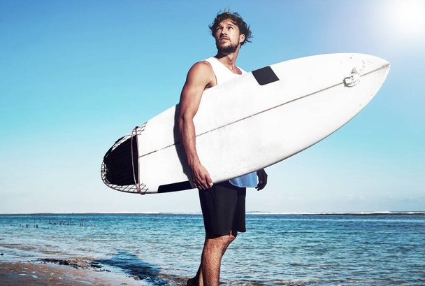Portrait of handsome hipster sunbathed fashion man model surfer wearing casual clothes going with surfboard on blue ocean and sky background - 写真・画像