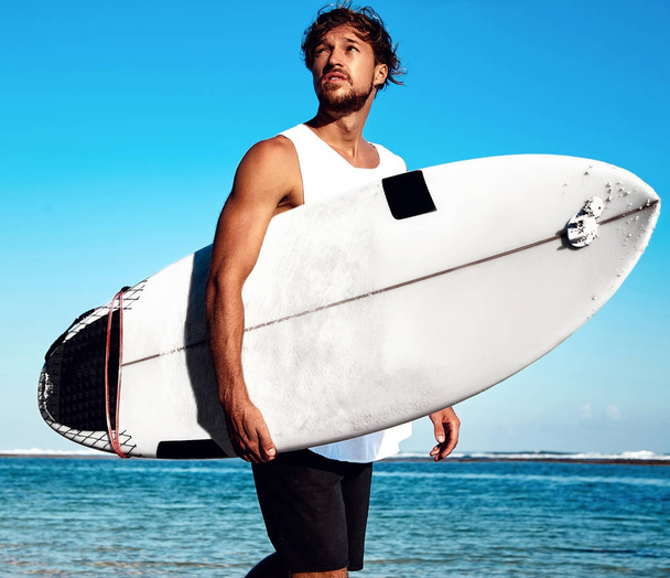 Portrait of handsome hipster sunbathed fashion man model surfer wearing casual clothes going with surfboard on blue ocean and sky background - Fotografie, Obrázek
