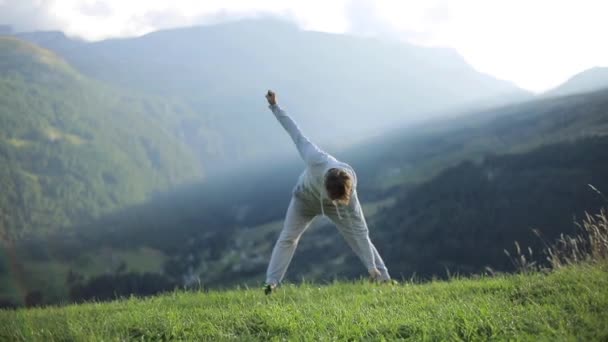 Man stands rotating hands cross touching toes warming up outdoors in mountain morning meadow slow motion. Man exercises works out wearing grey sport suit sneakers. Workout wellness fresh air lifestyle - Footage, Video