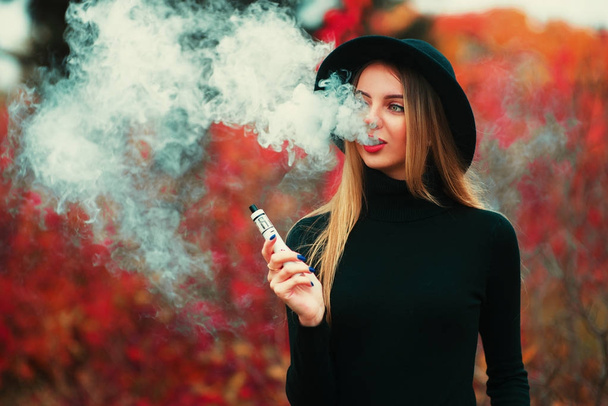 Pretty young hipster woman in black hat vape ecig, vaping device. - Photo, Image