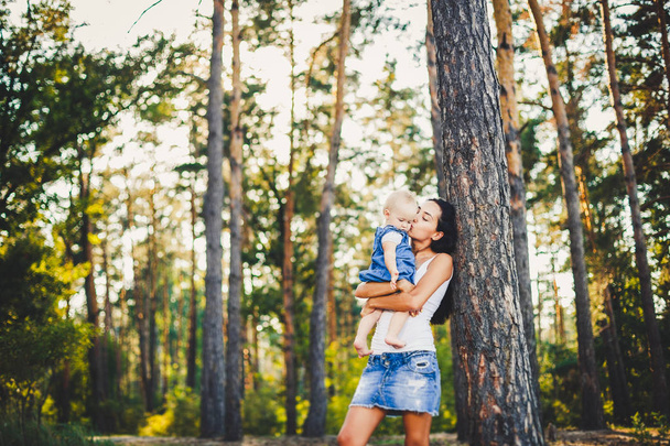 夏の公園の針葉樹の森の青い目をした金髪の娘の誕生の 1 年を保持している長いブルネットの髪を持つ若い美しいファッショナブルな母親. - 写真・画像