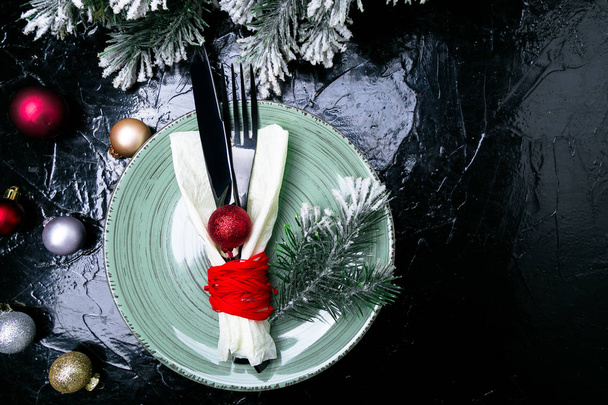 Vánoční prostírání stolu. Svátky na pozadí. Zelená deska, nůž a vidlička s vánoční výzdobou na černém stole. Pohled shora. - Fotografie, Obrázek