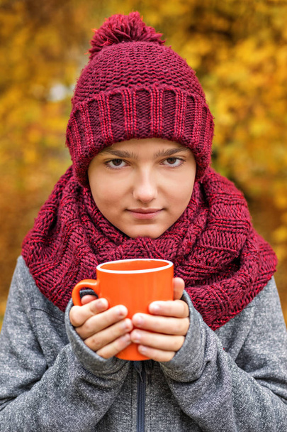  boy teenager with a cup of tea coffee in a voluminous large cozy scarf and cap of burgundy color - Foto, imagen