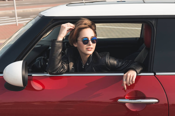 Beautiful girl in leather jacket and sun glasses is looking away while sitting in her car - Photo, Image