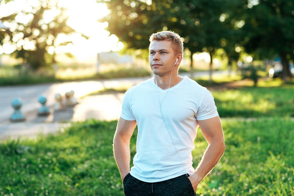 Man trainer in headphones. In a white T-shirt. Happy smiling. Listens to music, confident look. Summer lifestyle, motivation is strong. Outdoors in the city. - Valokuva, kuva