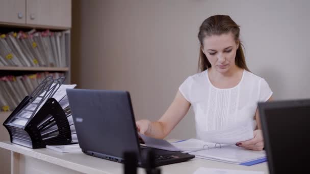 fiatal nő egy könyvelő irodában egy asztalnál ül, és ellenőrzése papírok, hümmögés mappában munkanap folyamán - Felvétel, videó